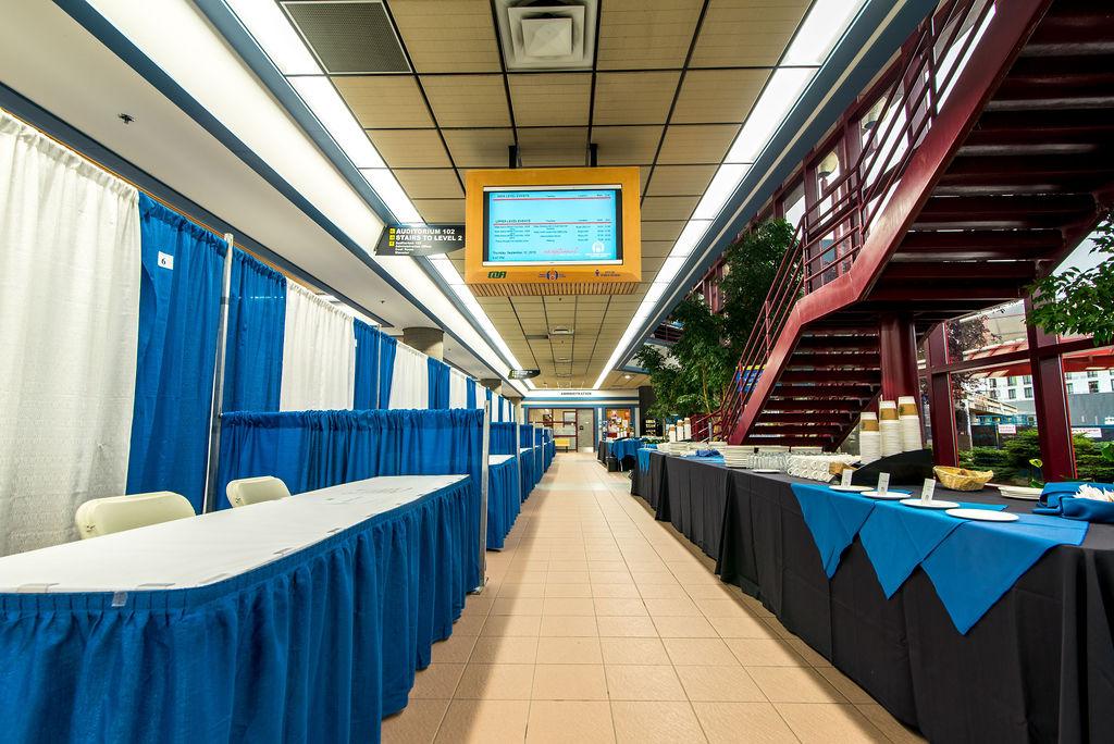 Lower area of Prince George Conference and Civic Centre. Area is set up for a trade show, with exhibition booths, skirted tables and folding chairs, and display monitor.